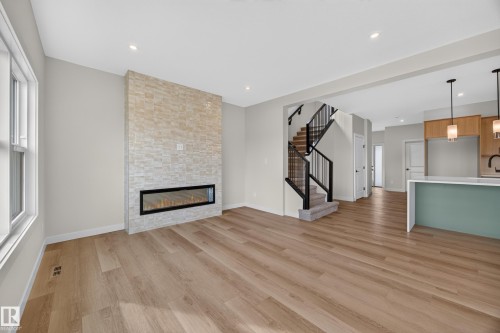 Spacious room featuring wood-finish flooring, a stone-clad fireplace with a linear insert, recessed lighting, a staircase with black metal balusters, and a kitchen island with a light-colored countertop - 205 Hampshire, Sherwood Park, AB 