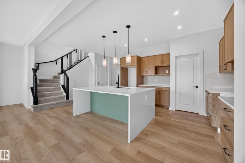 White waterfall-edge kitchen island, wood-finish flooring, recessed lighting, wood cabinetry, and a carpeted staircase with dark railings - 205 Hampshire, Sherwood Park, AB 