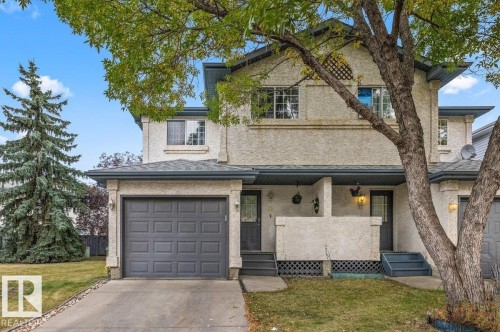 Stucco exterior with a dark gray garage door, paved driveway, and mature landscaping - #63 501 Youville Dr E Nw, Edmonton, AB 