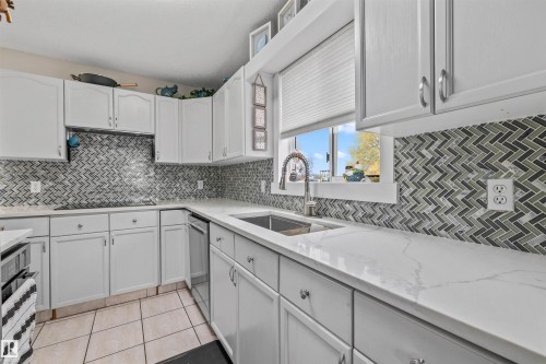 White cabinetry with stainless steel hardware, herringbone tile backsplash, white countertops with veining, and a stainless steel sink with a pull-down faucet - #63 501 Youville Dr E Nw, Edmonton, AB 