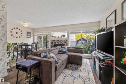 Open-concept living area with wood-finish flooring, a contemporary fireplace with a decorative tile surround, and abundant natural light from large windows - #63 501 Youville Dr E Nw, Edmonton, AB 