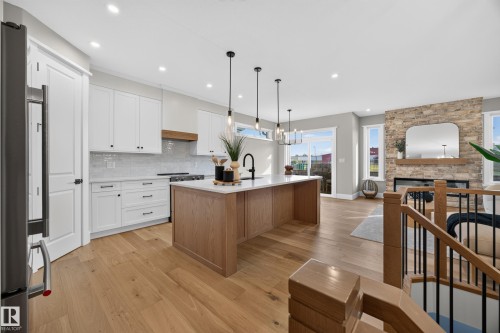 Modern kitchen featuring white cabinetry, light wood-finish flooring, a kitchen island with pendant lighting, and a stone-clad fireplace with a wood mantel - 209 Hampshire, Sherwood Park, AB 