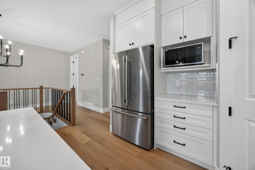 Integrated stainless steel refrigerator, built-in microwave, white shaker-style cabinetry with black hardware, light wood-finish flooring, and a contemporary stair railing with wood and black spindles - 209 Hampshire, Sherwood Park, AB 