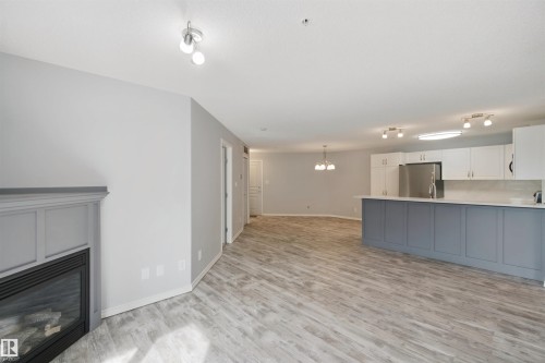 Wood-finish flooring throughout, featuring a built-in fireplace with a paneled surround, white upper cabinetry, a gray island with a countertop, and stainless steel appliances - #208 16035 132 St Nw, Edmonton, AB 
