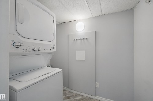 Stackable washer and dryer, light gray wall color, wood-finish flooring, and round wall-mounted light fixture - #208 16035 132 St Nw, Edmonton, AB 