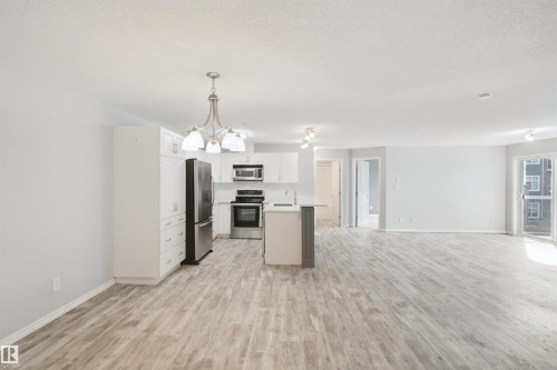 Bright open concept living area featuring wood-finish flooring, white cabinetry, stainless steel appliances, and a sliding glass patio door - #208 16035 132 St Nw, Edmonton, AB 