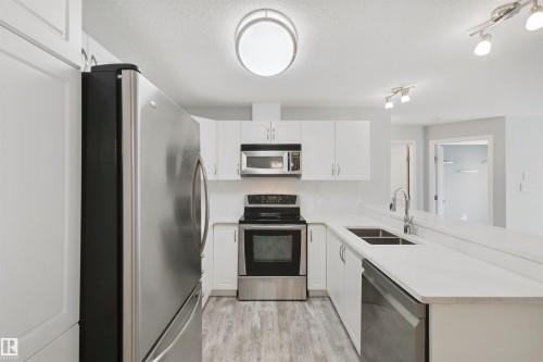 Kitchen featuring stainless steel appliances, white cabinetry, a double basin sink, and wood-finish flooring - #208 16035 132 St Nw, Edmonton, AB 