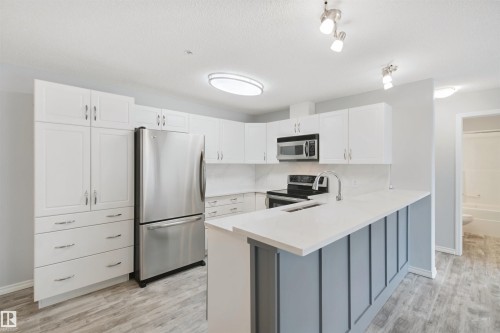 Kitchen featuring white cabinetry, stainless steel appliances, a white countertop, wood-finish flooring, and a breakfast bar - #208 16035 132 St Nw, Edmonton, AB 