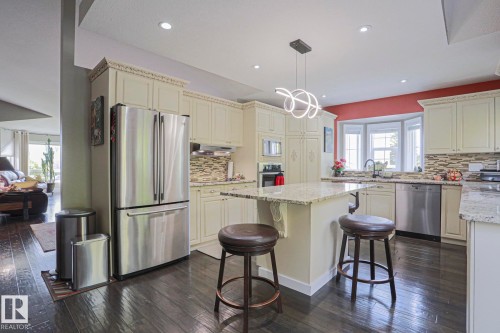 Gourmet kitchen featuring cream cabinetry with decorative molding, dark wood-finish flooring, stainless steel appliances, an eat-at island with light granite countertops, and a bay window - 3815 41 Street Nw, Edmonton, AB 