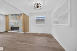 Wood-finish flooring, a contemporary electric fireplace with a decorative stone surround, an integrated wall shelving unit, a ceiling-mounted light fixture, and a window with white trim - 
