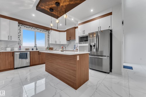 Modern kitchen featuring an island, white cabinetry with wood-finish accents, stainless steel appliances, and large format white tile flooring - 6502 42 Ave, Beaumont, AB 