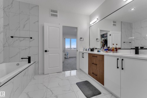 Bathroom featuring a built-in bathtub, full-height tile surround, dual vanity with wood-finish and white cabinetry, and matte black fixtures - 6502 42 Ave, Beaumont, AB 