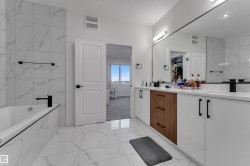Bathroom featuring a built-in bathtub, full-height tile surround, dual vanity with wood-finish and white cabinetry, and matte black fixtures - 