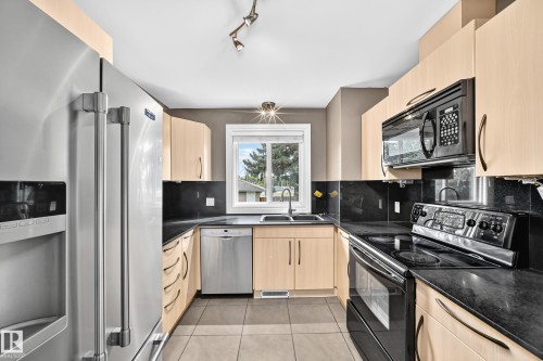 Kitchen featuring light wood-finish cabinetry, dark countertops, tile flooring, and stainless steel appliances including a refrigerator, dishwasher, and microwave - 10715 133 Street Nw, Edmonton, AB 