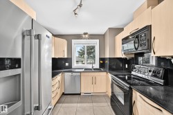 Kitchen featuring light wood-finish cabinetry, dark countertops, tile flooring, and stainless steel appliances including a refrigerator, dishwasher, and microwave - 