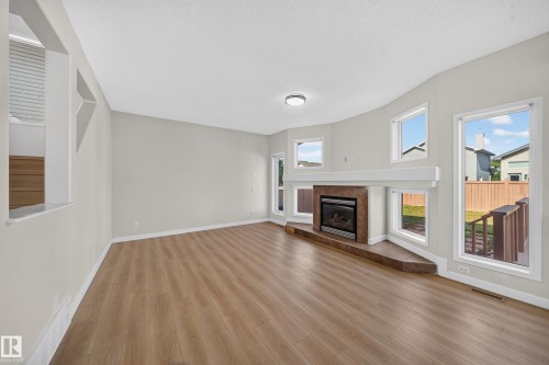 Spacious living area featuring wood-finish flooring, a corner fireplace with a tiled surround, multiple windows, and a built-in mantel - 7836 7 Ave Sw Sw, Edmonton, AB 