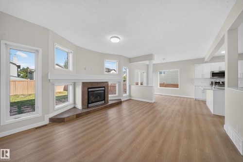 Spacious open-concept interior featuring light wood-finish flooring, a contemporary fireplace with a tiled surround, multiple large windows, and a modern kitchen with white cabinetry and stainless steel appliances - 7836 7 Ave Sw Sw, Edmonton, AB 
