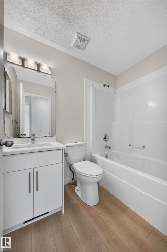 Bathroom featuring a single vanity with storage, a rectangular mirror, a bathtub with a shower, and wood-finish flooring - 7836 7 Ave Sw Sw, Edmonton, AB 