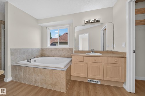 Soaking tub with tiled surround, wood-finish flooring, single vanity with integrated sink, wood-finish cabinetry, and a framed mirror with a three-light fixture - 7836 7 Ave Sw Sw, Edmonton, AB 