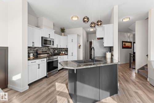 Kitchen featuring white cabinetry, stainless steel appliances, a dark backsplash, a kitchen island with a double basin sink, and wood-finish flooring - #5 4900 62 St, Beaumont, AB 