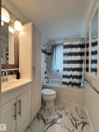 White vanity with integrated sink, modern wall sconces, white porcelain tile flooring with dark veining, built-in tub with white subway tile surround, and white storage cabinetry - 