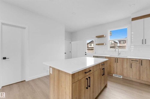 Kitchen featuring a central island with white countertop, wood-finish cabinetry with matte black hardware, white subway tile backsplash, and light wood-finish flooring - 523 Robinson Blvd, Leduc, AB 
