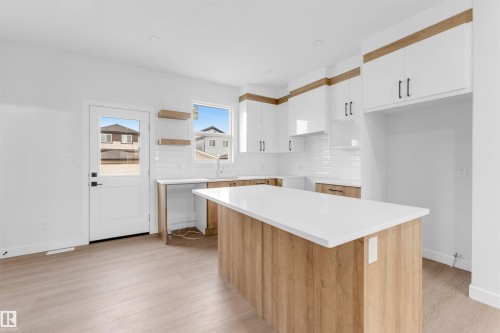 Modern kitchen featuring a central island with a white countertop, white upper cabinetry with wood-finish accents, wood-finish lower cabinetry, a white subway tile backsplash, and wood-finish flooring - 523 Robinson Blvd, Leduc, AB 