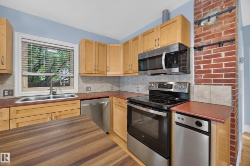 Kitchen featuring light wood cabinetry, stainless steel appliances, a double basin sink, tile backsplash, and a brick accent column - 9615 75 Avenue Nw, Edmonton, AB 