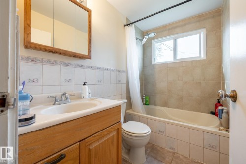 Bathroom featuring a single vanity with wood-finish cabinetry, a toilet, a bathtub with an integrated shower, a window, and wall-mounted tile - 10972 157 Street Nw, Edmonton, AB 