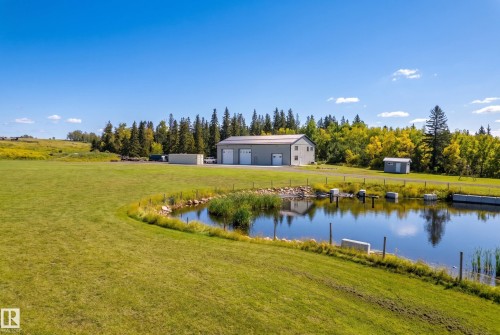 Expansive green space featuring a water body, a large two-story outbuilding with multiple garage doors, and additional storage structures - 49279 Rge Rd 240, Rural Leduc County, AB 
