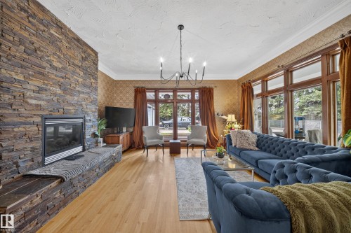 Stacked stone accent wall with an integrated fireplace and hearth, wood-finish flooring, a chandelier, and expansive windows with dark wood frames - 719 65 Ave Nw, Edmonton, AB 