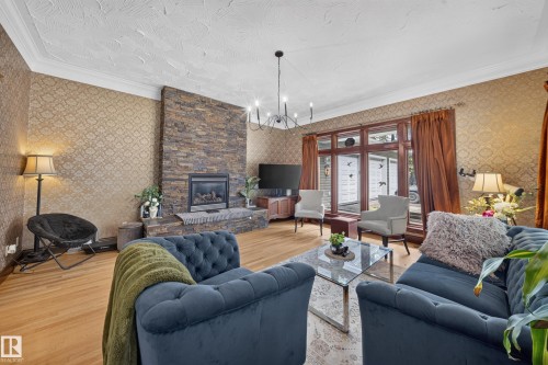 Stacked stone fireplace with a built-in hearth, wood-finish flooring, patterned wall covering, a large window with wood trim, and a chandelier lighting fixture - 719 65 Ave Nw, Edmonton, AB 