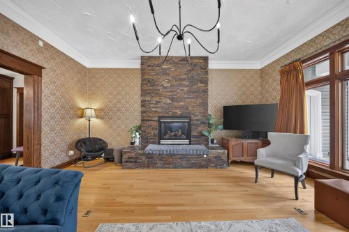 Stacked stone fireplace with a raised hearth, wood-finish flooring, decorative wall covering, crown molding, and a multi-arm chandelier - 719 65 Ave Nw, Edmonton, AB 