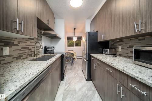 Galley kitchen featuring wood-look cabinetry, granite-style countertops, and a full-height tile backsplash - #202 10615 156 St Nw, Edmonton, AB 