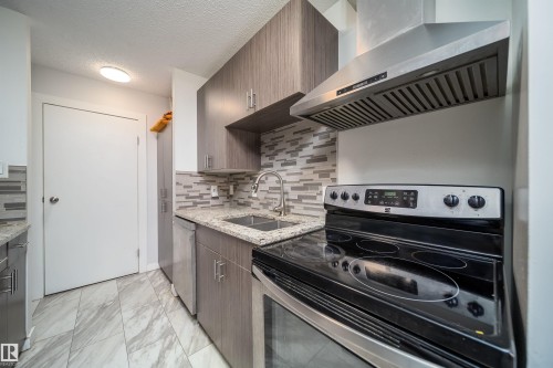 Kitchen featuring wood-finish cabinetry, speckled countertops, stainless steel range hood, glass-top electric range, and a mosaic tile backsplash - #202 10615 156 St Nw, Edmonton, AB 