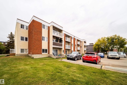 Multi-story building featuring brick and stucco exterior, multiple balconies with white railings, ground-level patios, and an asphalt parking area with designated spaces - #202 10615 156 St Nw, Edmonton, AB 
