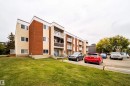 Multi-story building featuring brick and stucco exterior, multiple balconies with white railings, ground-level patios, and an asphalt parking area with designated spaces - #202 10615 156 St Nw, Edmonton, AB 