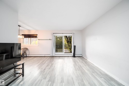 Expansive room featuring light wood-finish flooring, a sliding glass door leading to a balcony, and a prominent window - #202 10615 156 St Nw, Edmonton, AB 