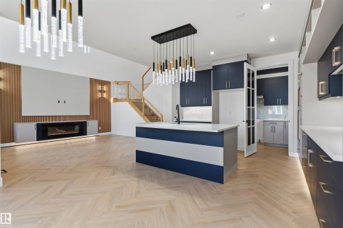 Herringbone wood-finish flooring, two-tone kitchen island with a built-in sink, dark blue cabinetry with gold-tone hardware, contemporary linear fireplace, and a wood-slat accent wall - 2720 63 Avenue, Rural Leduc County, AB 