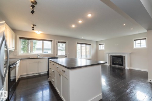 Open concept living area featuring dark wood-finish flooring, a white kitchen island with a dark countertop, white shaker cabinetry with stainless steel hardware, and a gas fireplace with a white mantel - 55 Michigan Street, Devon, AB 