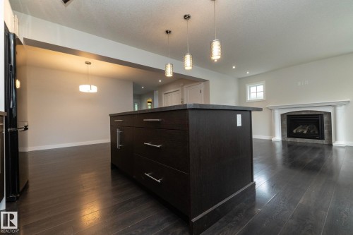 Dark wood-finish flooring, central island with dark cabinetry, three pendant lights, recessed lighting, and a fireplace with a decorative mantel and hearth - 53 Michigan Street, Devon, AB 