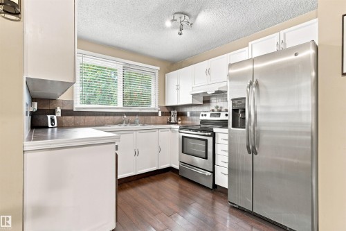 Kitchen featuring white cabinetry, stainless steel appliances, wood-finish flooring, and a window with blinds - 12 Hughes Road Nw, Edmonton, AB 