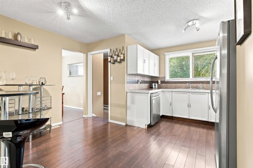 Kitchen featuring wood-finish flooring, white cabinetry, a stainless steel dishwasher, a stainless steel refrigerator, and a window with blinds - 12 Hughes Road Nw, Edmonton, AB 