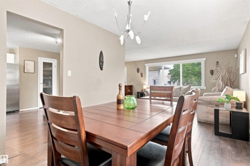 Open concept living and dining areas featuring wood-finish flooring, neutral wall tones, a contemporary chandelier, and a large window with horizontal blinds - 12 Hughes Road Nw, Edmonton, AB 