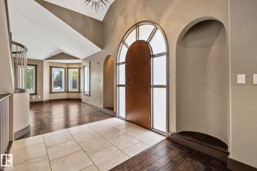Arched entry door with sidelights, featuring tile and wood-finish flooring, a curved staircase with metal railings, and bay windows with plantation shutters - 10 Eldorado Drive, St. Albert, AB 