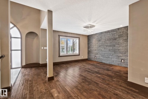 Stone accent wall with integrated floor lighting, wood-finish flooring, square crystal chandelier, arched entryway with frosted glass panel, and window with plantation shutters - 10 Eldorado Drive, St. Albert, AB 