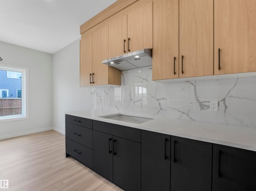 Kitchen featuring light wood-finish upper cabinetry, dark lower cabinetry, a stainless steel range hood, stone-finish countertops, and a full-height stone-finish backsplash - 11 Dalquist Bay, Leduc, AB 