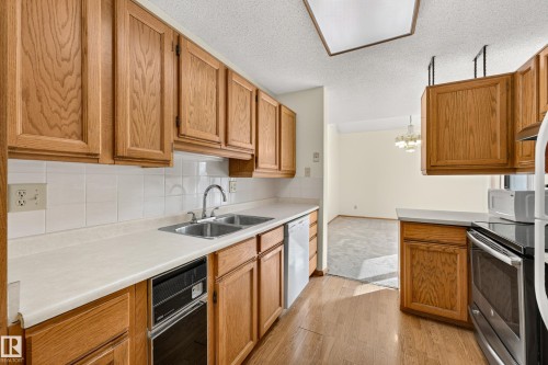 Kitchen featuring wood-finish cabinetry, white countertops, a double-basin stainless steel sink, and white tile backsplash - #23 500 Lessard Dr Nw, Edmonton, AB 