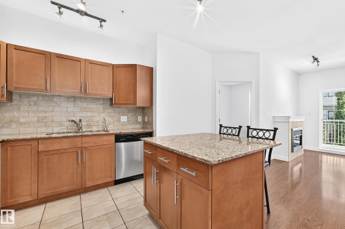 Kitchen featuring wood cabinetry, a granite countertop island with seating, a tile backsplash, and stainless steel appliances - #307 1003 Gault Bv Nw, Edmonton, AB 