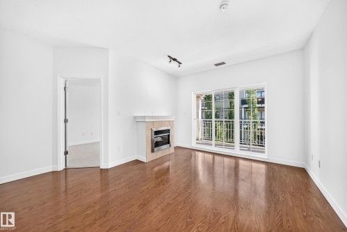 Wood-finish flooring throughout, bright white walls, fireplace with stone surround, track lighting, and a sliding glass door opening to a balcony - #307 1003 Gault Bv Nw, Edmonton, AB 